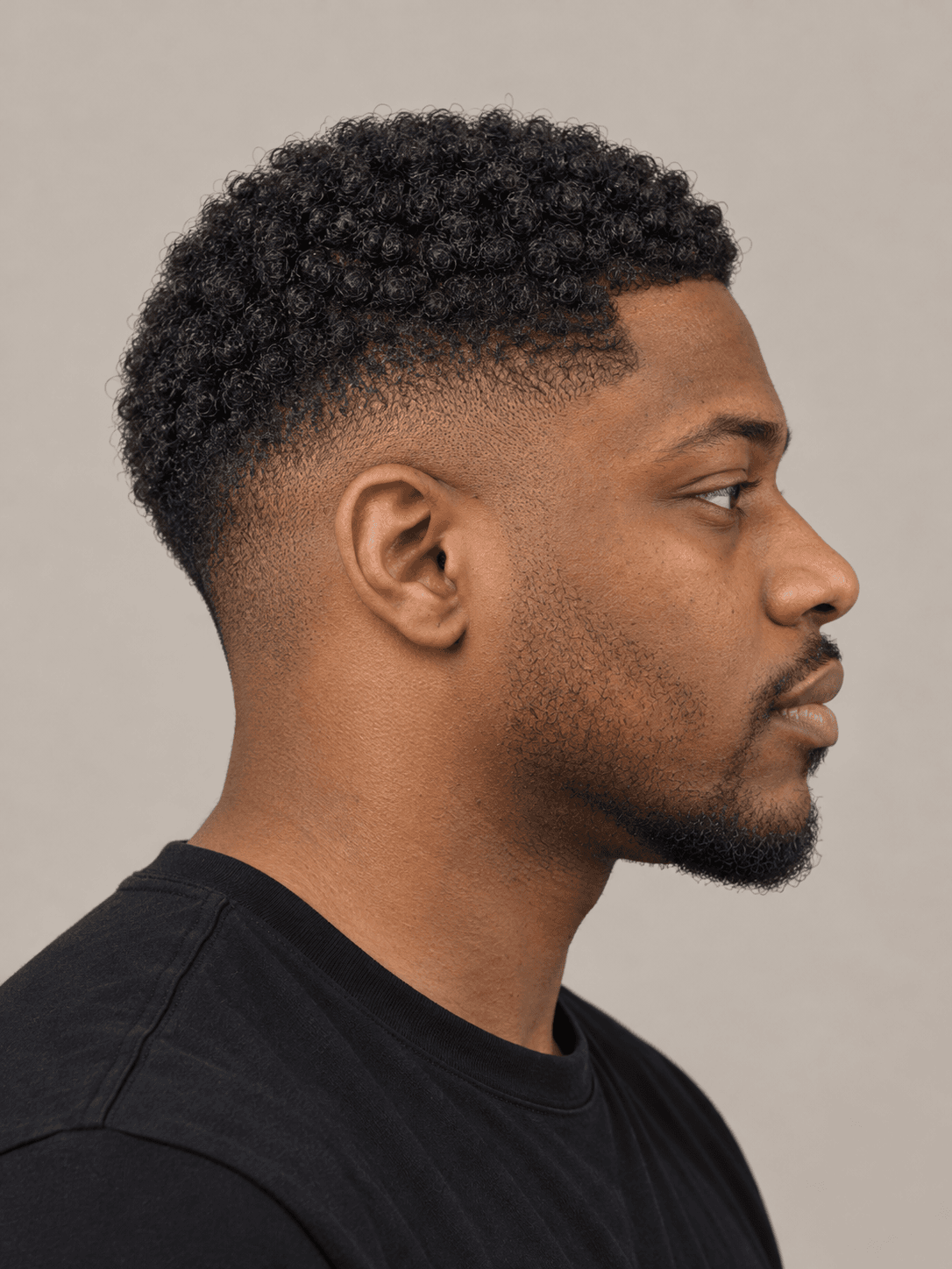 Low fade hairstyle with a curly top on a man with black coily hair and a beard, side view
