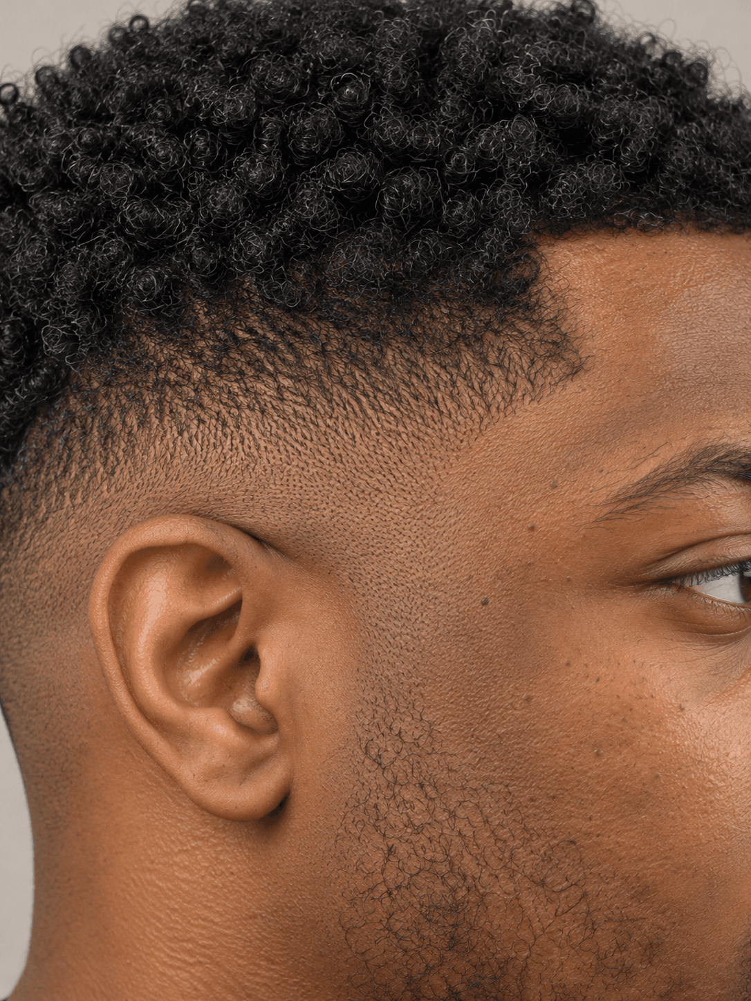 Low fade hairstyle with a curly top on a man with black coily hair and a beard, close-up of the fade from ear to temple